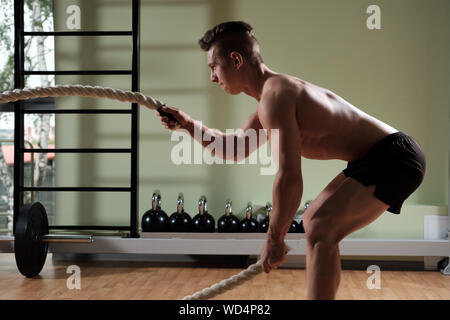 Muscular man with battle rope doing exercise in functional training gym. Profile view. Stock Photo
