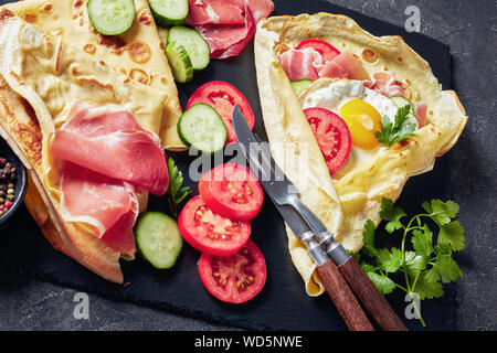 delicious breakfast of Crepes with fried egg, Jamon slices, fresh tomatoes and cucumber salad fillings on a black slate tray with ingredients, view fr