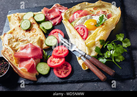 delicious breakfast of Crepes with fried egg, Jamon slices, fresh tomatoes and cucumber fillings on a black slate tray with ingredients, view from abo Stock Photo