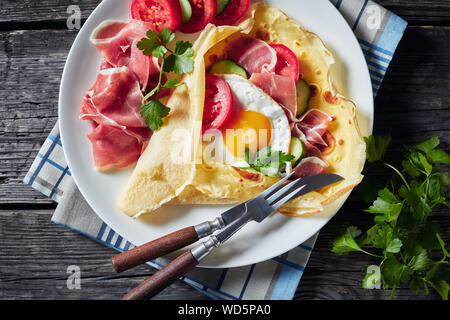Crepes with fried egg, Jamon slices, fresh tomatoes and cucumber fillings on a white plate on an old wooden table, view from above, flatlay, close-up Stock Photo