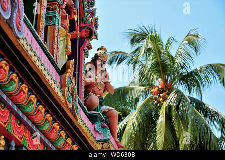 Stone statue of Hindu Indian deity in prayer on side of colourful temple wall with palm tree in soft focus background Stock Photo