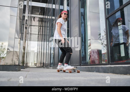 Photo of sporty brunette woman riding skateboard on street among modern buildings on summer day Stock Photo