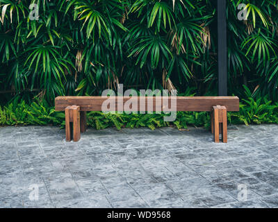 Rustic solid railway sleeper wooden bench on concrete floor on tropical green bush background. Stock Photo