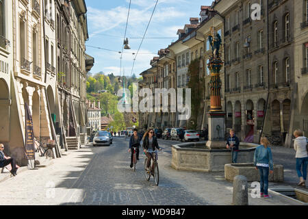 Couple cycling along street of Gerechtigkeitsgasse and past  Gerechtigkeitsbrunnen fountain statue. Old Town, Bern, Switzerland Stock Photo