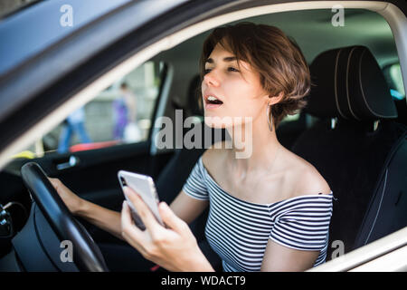 Closeup portrait, young woman driving in black car and checking her phone, annoyed by navigation gps system or bad text message or email, isolated out Stock Photo