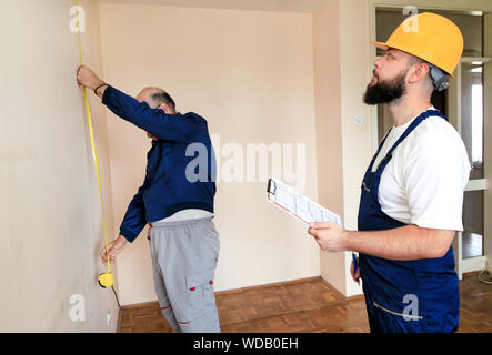Engineer, contractor and project manager with his colleague, construction worker, handyman and builder is measuring wall of room using measure tape. Stock Photo