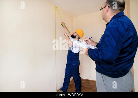 Engineer, contractor and project manager with his colleague, construction worker, handyman and builder is measuring wall of room using measure tape. Stock Photo