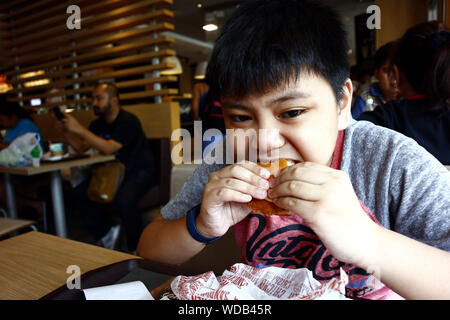 SAN JUAN, METRO MANILA, PHILIPPINES – AUGUST 21, 2019: Young Asian boy eats a hamburger inside a fast food restaurant. Stock Photo