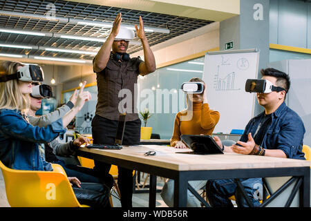 Testing games. Young African man wearing virtual reality headset and gesturing while standing at the desk in creative office together with four multie Stock Photo