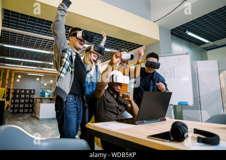Young cheerful team of young testers celebrating victory in a project while using VR glasses. Stock Photo