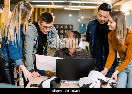 Expressive young multiethnical people working together in modern office. Cheerful African man sitting at the table with laptop Stock Photo
