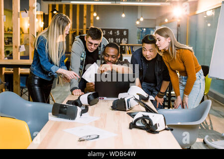 Group of Adult Hipsters coworkers Using Modern Gadgets. Four Business friends, smiling and gesturing while their African friend works on laptop at the Stock Photo