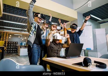 Supportive group of young diverse multiracial business people wearing vr goggles celebrating their victory in creative modern loft office. Stock Photo