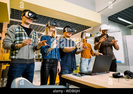 Five multiethnical excited smiling businesspeople wearing virtual reality glasses, interacting with digital interface while standing near table in mod Stock Photo