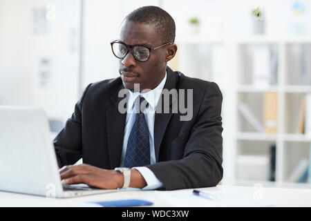 Concentrated young African businessman wearing eyeglasses and formal wear working on laptop computer at the workplace Stock Photo