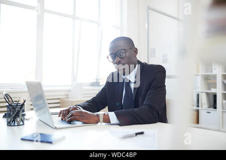 Portrait of African young businessman in eyeglasses wearing black suit sitting at the table and looking at camera while typing on laptop Stock Photo