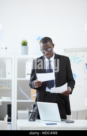 Serious African manager in suit standing and concentrating on reading of documents while working at the office Stock Photo