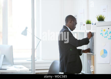 Serious African businessman in suit standing near the office closet and getting the document from folder while working at modern office Stock Photo