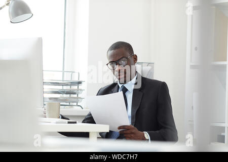 Portrait of confident African businessman wearing eyeglasses holding document and looking at camera while drinking coffee at office Stock Photo