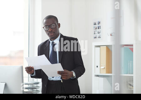 Portrait of African young office worker standing in formal wear holding documents and looking at camera at modern office Stock Photo