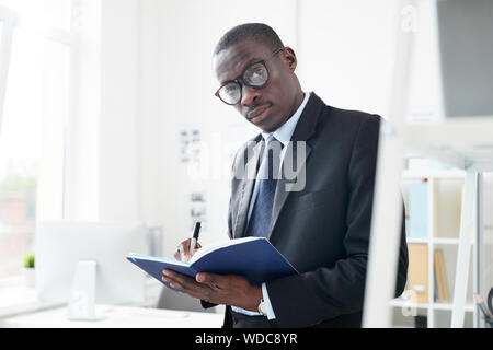 Portrait of African young businessman in eyeglasses wearing formal wear writing his plans in notepad while standing at office Stock Photo