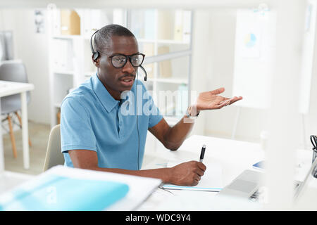 African young man in casual clothing sitting at the table wearing headphones and explaining something on the phone he working in a call center Stock Photo