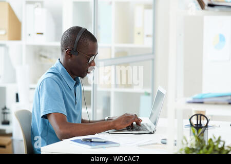 Serious African programmer in casual clothing wearing headphones and concentrating on his work on laptop computer at office Stock Photo
