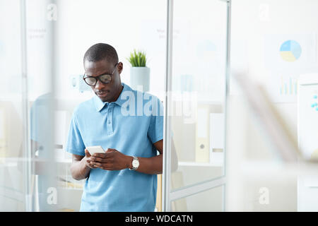 Young African office worker in casual clothing standing at office and typing a message on mobile phone Stock Photo