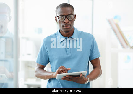 Portrait of African young man in eyeglasses and in blue shirt looking at camera while using digital tablet Stock Photo