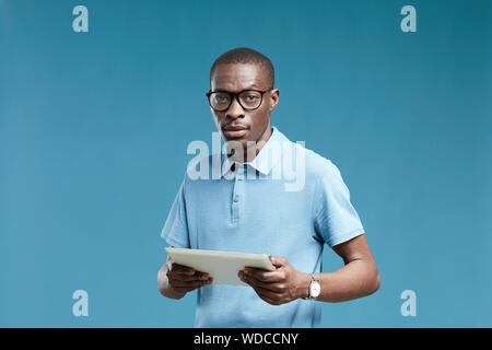 Portrait of young African man in casual clothing and in eyeglasses holding digital tablet and looking at camera over blue background Stock Photo