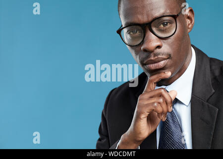 Portrait of African young office worker in eyeglasses and suit looking thoughtfully over blue background Stock Photo