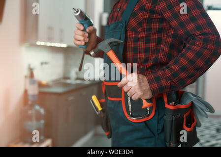 Cropped shot of man in checkered shirt wearing tool kit standing in the kitchen, holding a hammer and a drill in his hands. Horizontal shot Stock Photo