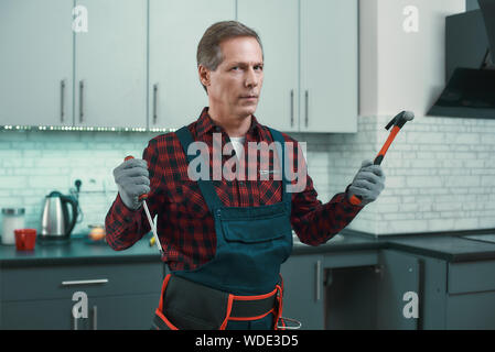 Portrait of serious caucasian repairman in uniform standing in the kitchen and looking at the camera. He holds a screwdriver and a hammer in orer to start working. Repair service concept Stock Photo