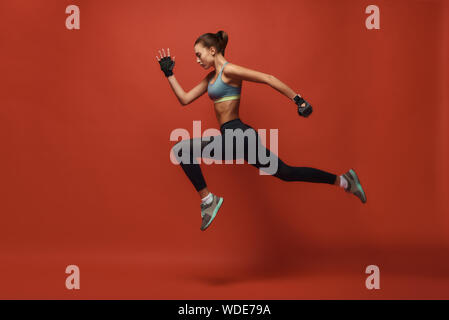 Full-length portrait of young girl in sportswear jumping isolated over red background. Fitness and work out concept. Horizontal shot. Side view Stock Photo