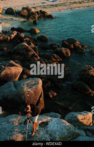 Vertical shot of a photographer setting up his camera on a rock near the water Stock Photo