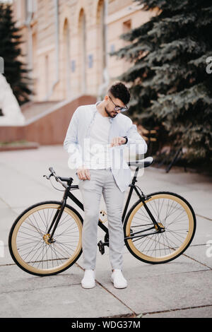 Handsome young businessman looking at watch while standing near bicycle outdoors on sunny day Stock Photo