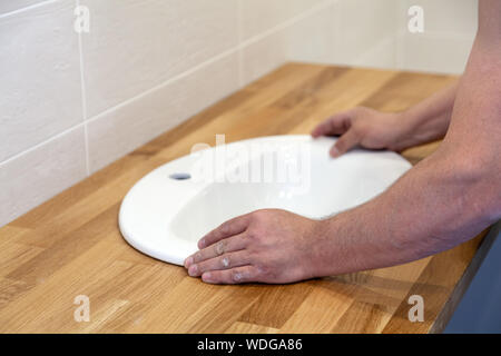 Closeup of hands of professional plumber worker installs white oval ceramic sink on wooden tabletop in bathroom with beige tile Stock Photo