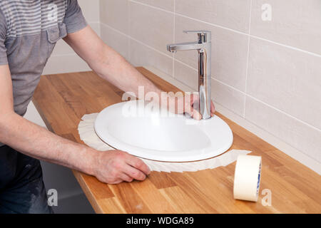 Closeup hands of a professional plumber worker installs a white oval ceramic sink on a wooden tabletop in the bathroom with beige tile, paste over the Stock Photo