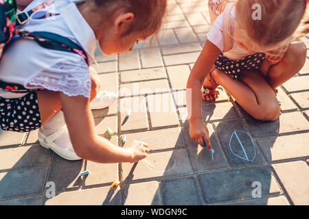 Happy little girls wearing backpacks and drawing with chalk outdoors primary school. Kids having fun after classes. Back to school Stock Photo