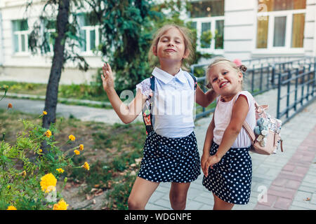 Two sisters pupils with backpacks grimacing outdoors primary school after classes. Little girls having fun. Happy kids smiling Stock Photo