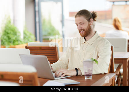 Young bearded man communicating online on his laptop and smiling while sitting at the table in cafe Stock Photo