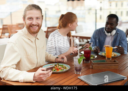 Portrait of bearded young man with mobile phone smiling at camera while have a lunch together with his friends in the background in cafe Stock Photo