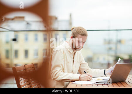 Young bearded man sitting at the table in front of laptop and making notes in document in outdoor coffee shop Stock Photo