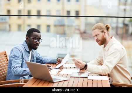 Two multiethnic business colleagues sitting at the table using laptop reading document and discussing it together in team in outdoor cafe Stock Photo