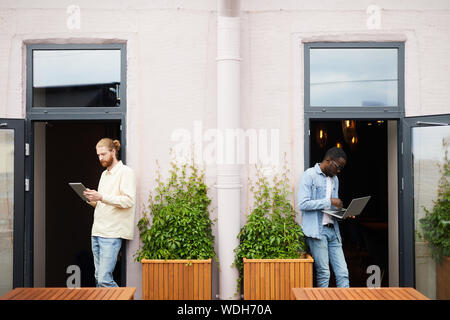 Young bearded man using digital tablet with other African young man working on laptop computer they standing near the entrance in cafe Stock Photo