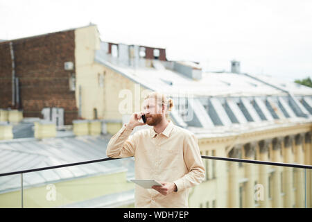 Bearded young man holding digital tablet and talking on mobile phone while standing outdoors with beautiful building in the background Stock Photo
