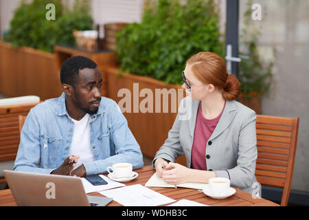 Young businesswoman has a business interview with African young man they sitting at the table with laptop and documents and talking in cafe Stock Photo