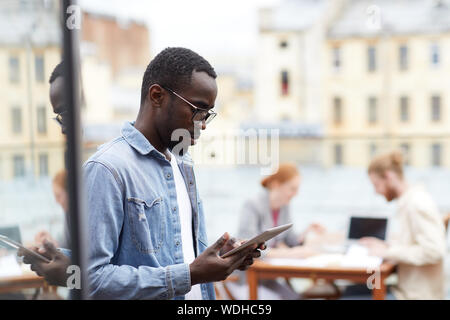 Serious young African man in eyeglasses communicating online using digital tablet while standing outdoors near the cafe Stock Photo