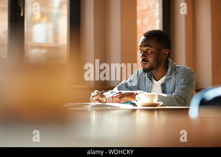 Pensive African man sitting at the table with cup of coffee making notes in notepad and thinking over new ideas in cafe Stock Photo