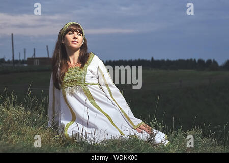 beautiful dark-haired calm girl in White Russian national costume sitting on the grass, background, field, sky, evening Stock Photo